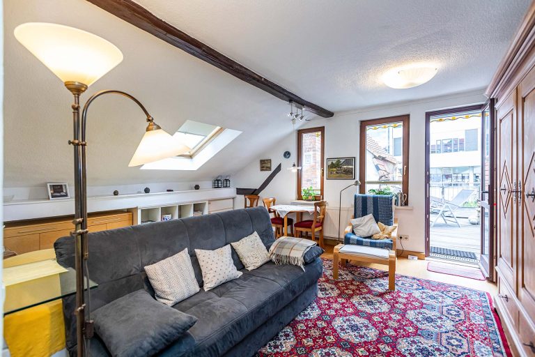 Wohn- und Schlafbereich der Ferienwohnung Schwing mit einem Dachfenster, einem Bett, einem anthrazitfarbenen Sofa, Holztisch, Holzschrank, Sessel, Tisch, Stühlen und Blick auf die Dachterrasse.